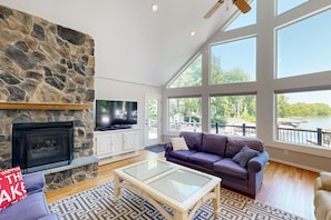 A spacious living room with a stone fireplace, a television, a large window with a lake view, and a leather sofa. - Exquisite Dog-friendly Home With Kayaks, Pool Table, hot Tub, & Washer/dryer (Bumpass)