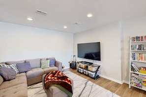 A living room with a sectional sofa, a flat-screen TV, a bookshelf, and a patterned rug. - Exquisite Dog-friendly Home With Kayaks, Pool Table, hot Tub, & Washer/dryer (Bumpass)