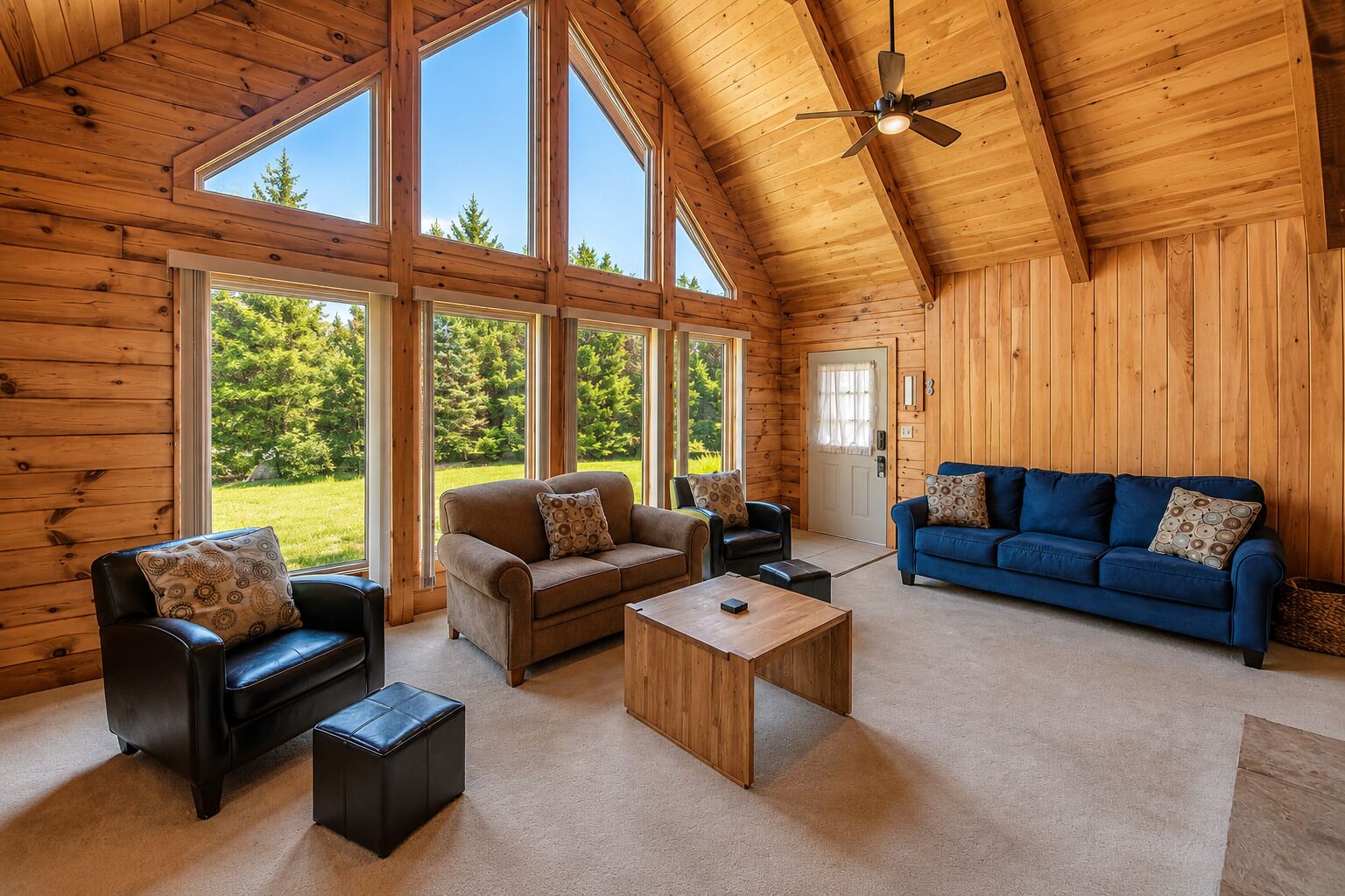 Interior de una cabaña de madera con techo alto, ventanas amplias, un sofá azul, un sofá marrón, un sillón de cuero negro y una mesa de centro de madera.