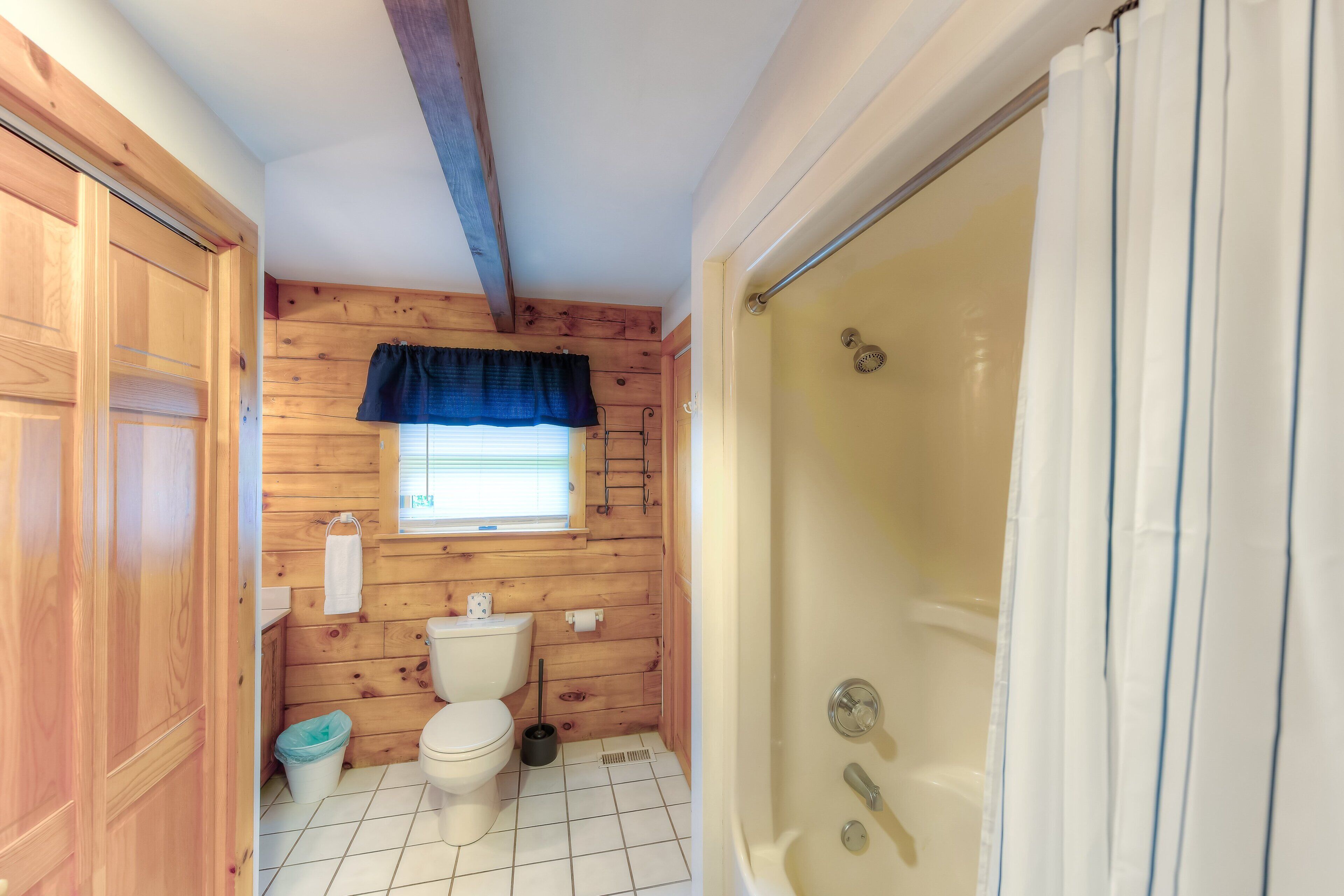 A bathroom with wooden walls, a white toilet, a shower, and a window with a blue curtain.