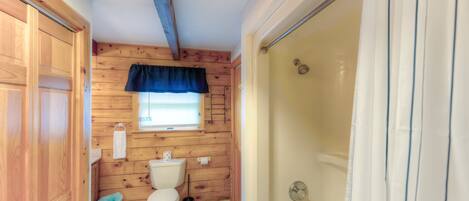A bathroom with wooden walls, a white toilet, a shower, and a window with a blue curtain.