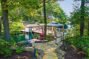 A wooden deck with a gazebo, a dock, and a small building near a body of water surrounded by trees.