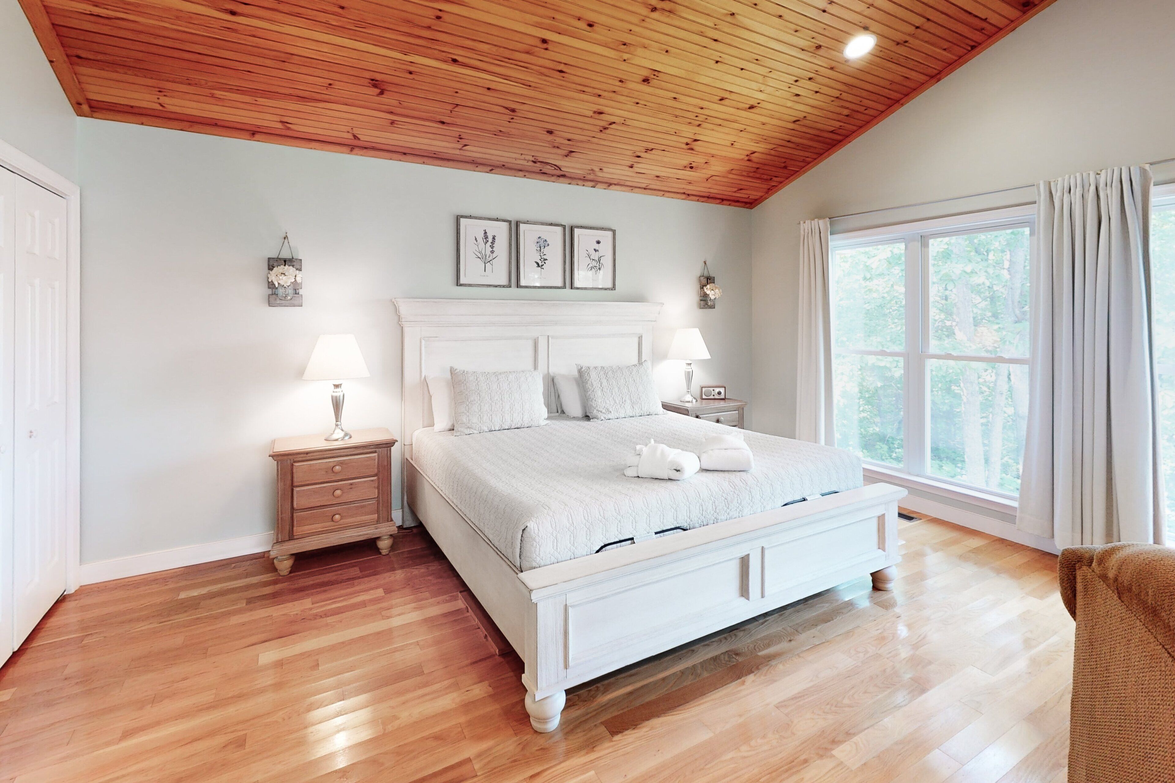 A bedroom with a wooden ceiling, a large bed with white bedding, two bedside tables with lamps, and a window with curtains.