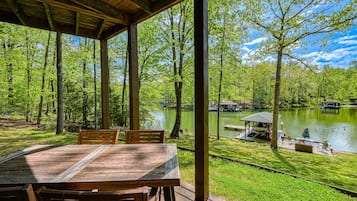 A covered outdoor area with a wooden table and chairs, overlooking a lake with a dock and a gazebo.