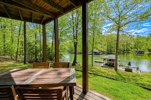 A covered outdoor area with a wooden table and chairs, overlooking a lake with a dock and a gazebo.