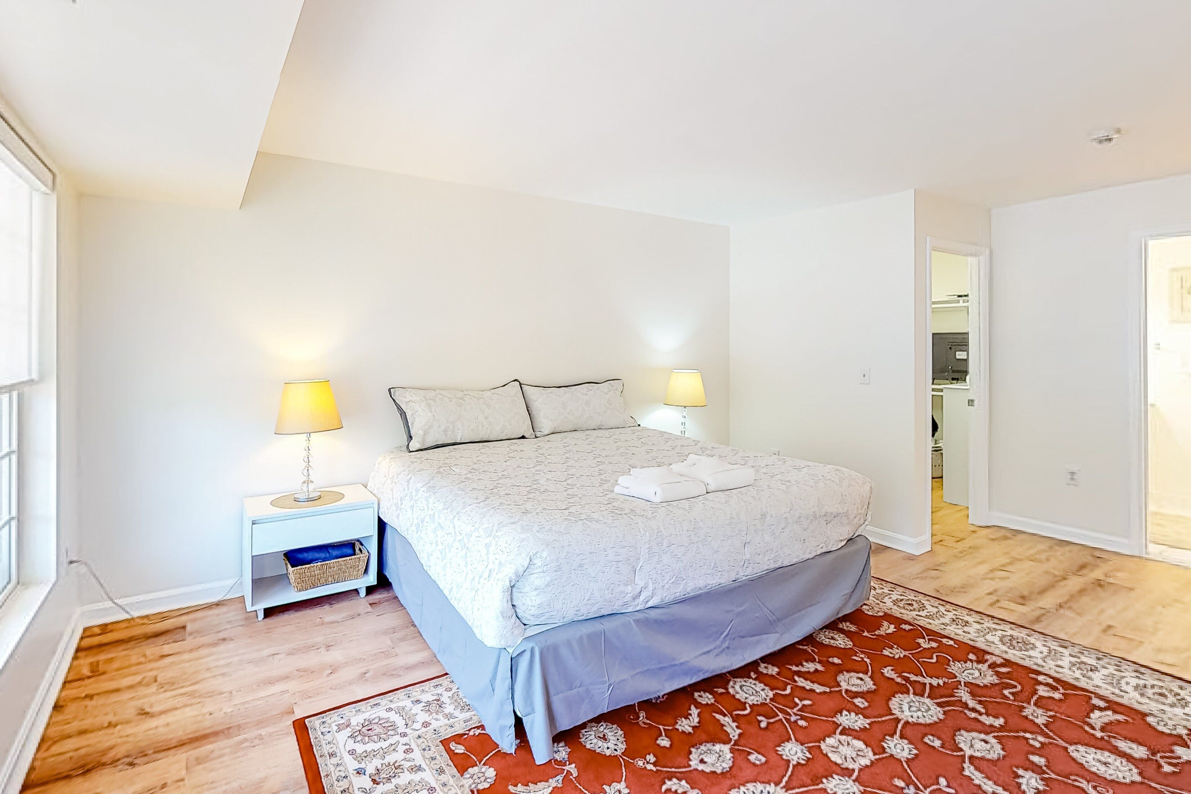 A bedroom with a bed, bedside lamps, a nightstand, a patterned rug, and a window.