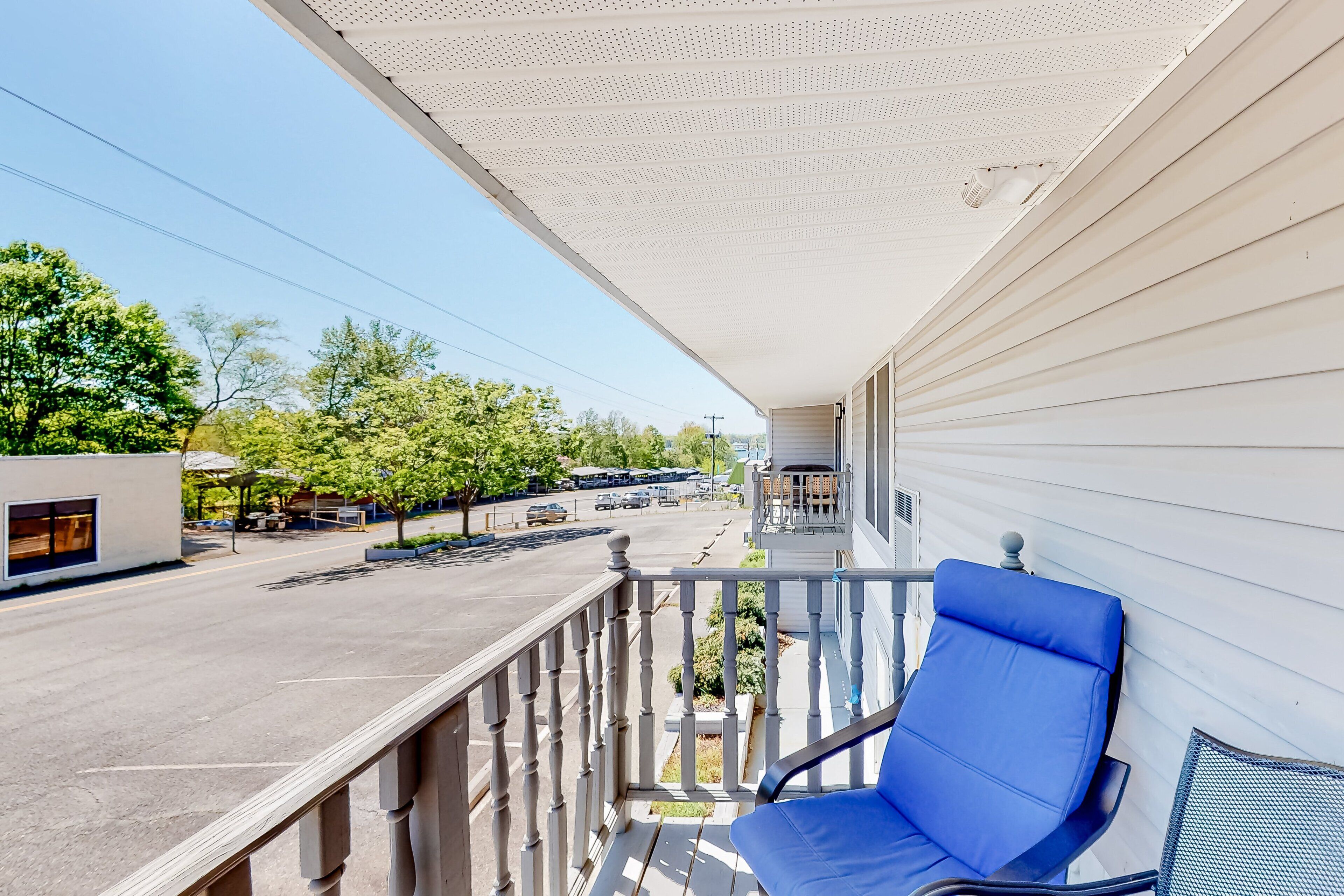 A balcony with a blue chair, a white ceiling, and a view of a street with parked cars and trees.