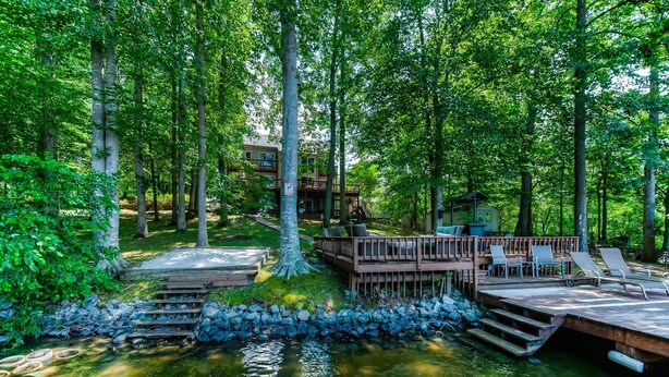 A wooden deck with lounge chairs, a small building, and a stone wall by a body of water surrounded by trees. - Spacious Lake Lodge With a Private Dock, Wraparound Decks & Foosball (Mineral)