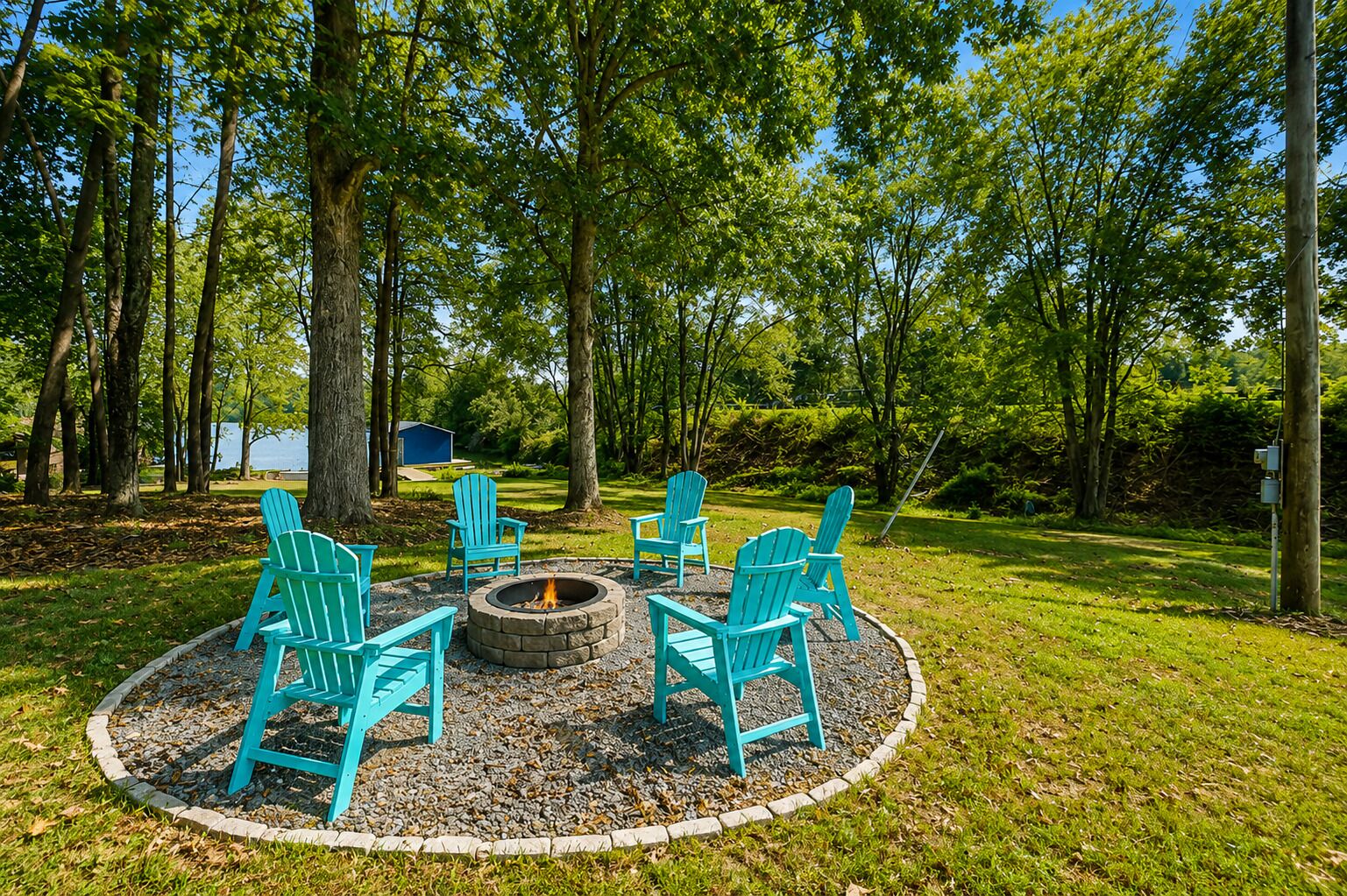 Un espace circulaire aménagé avec des chaises disposées autour d’un foyer, le tout bordé de gravier, dans un cadre verdoyant évoquant un parc, avec des arbres et une étendue d’eau en arrière-plan.