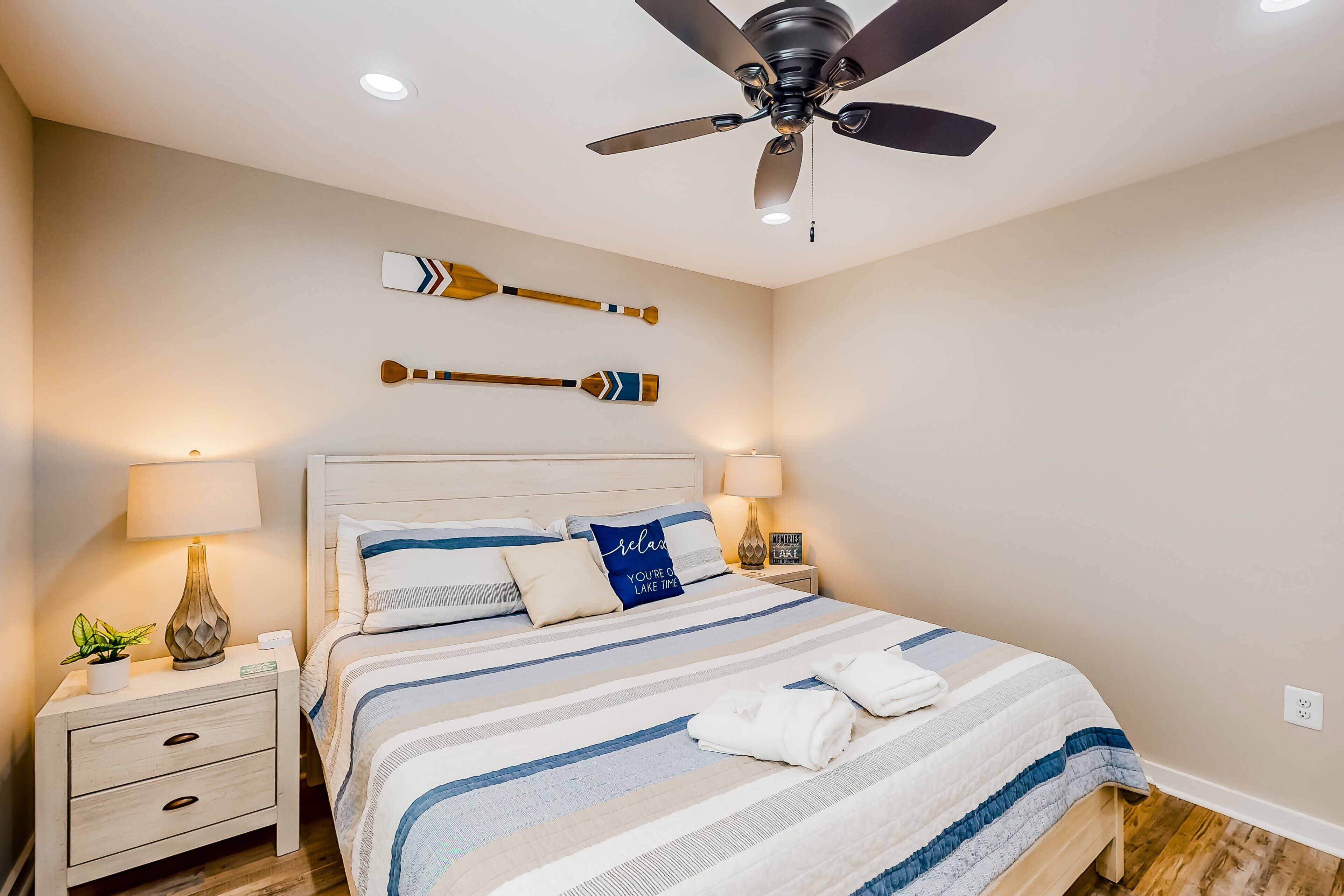 A bedroom with a bed, nightstands, lamps, a ceiling fan, and wall-mounted decorative items.