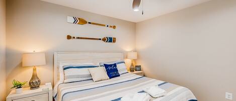 A bedroom with a bed, nightstands, lamps, a ceiling fan, and wall-mounted decorative items.