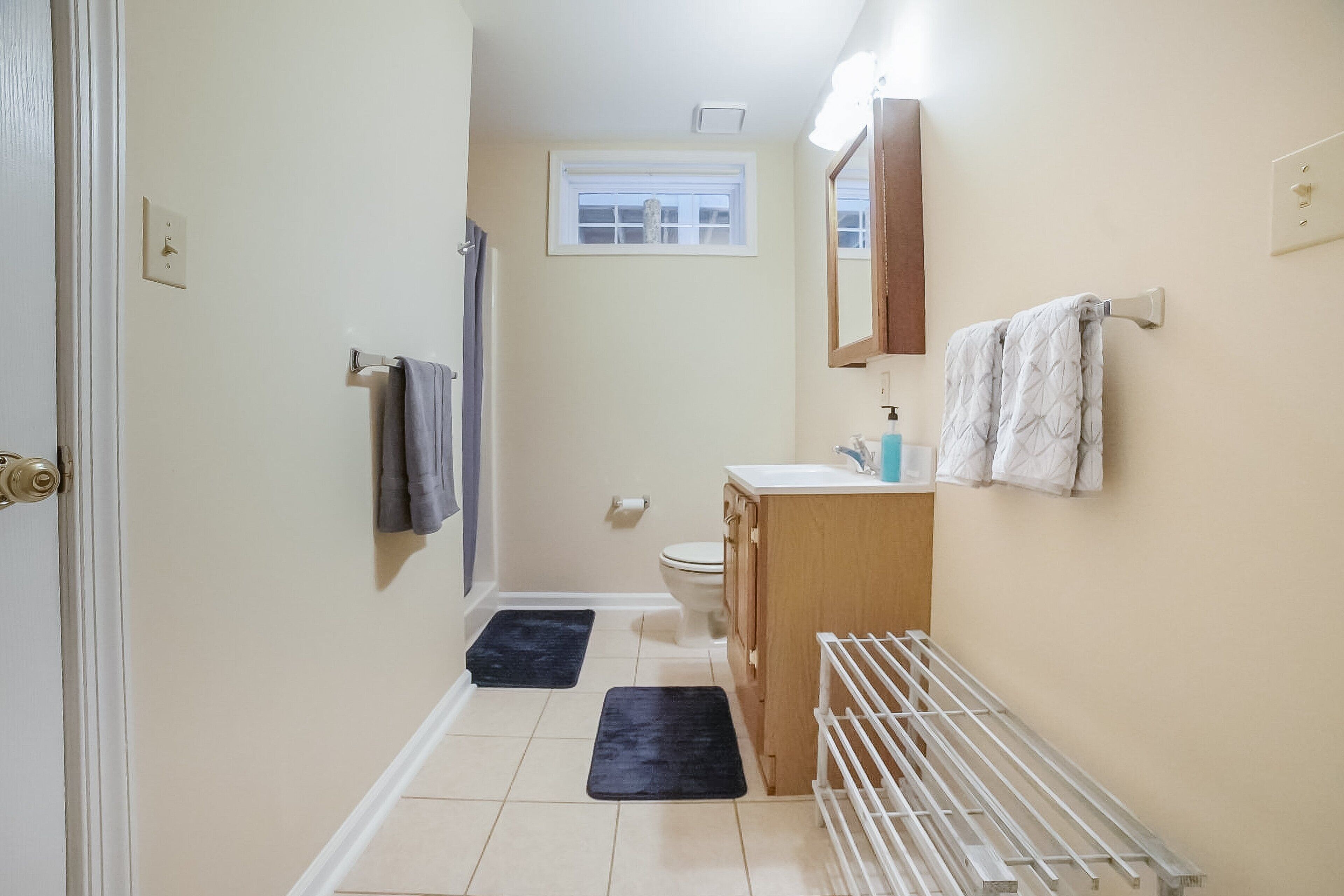 A narrow bathroom with a toilet, sink, and mirror. There are two mats on the floor and a towel rack on the wall.