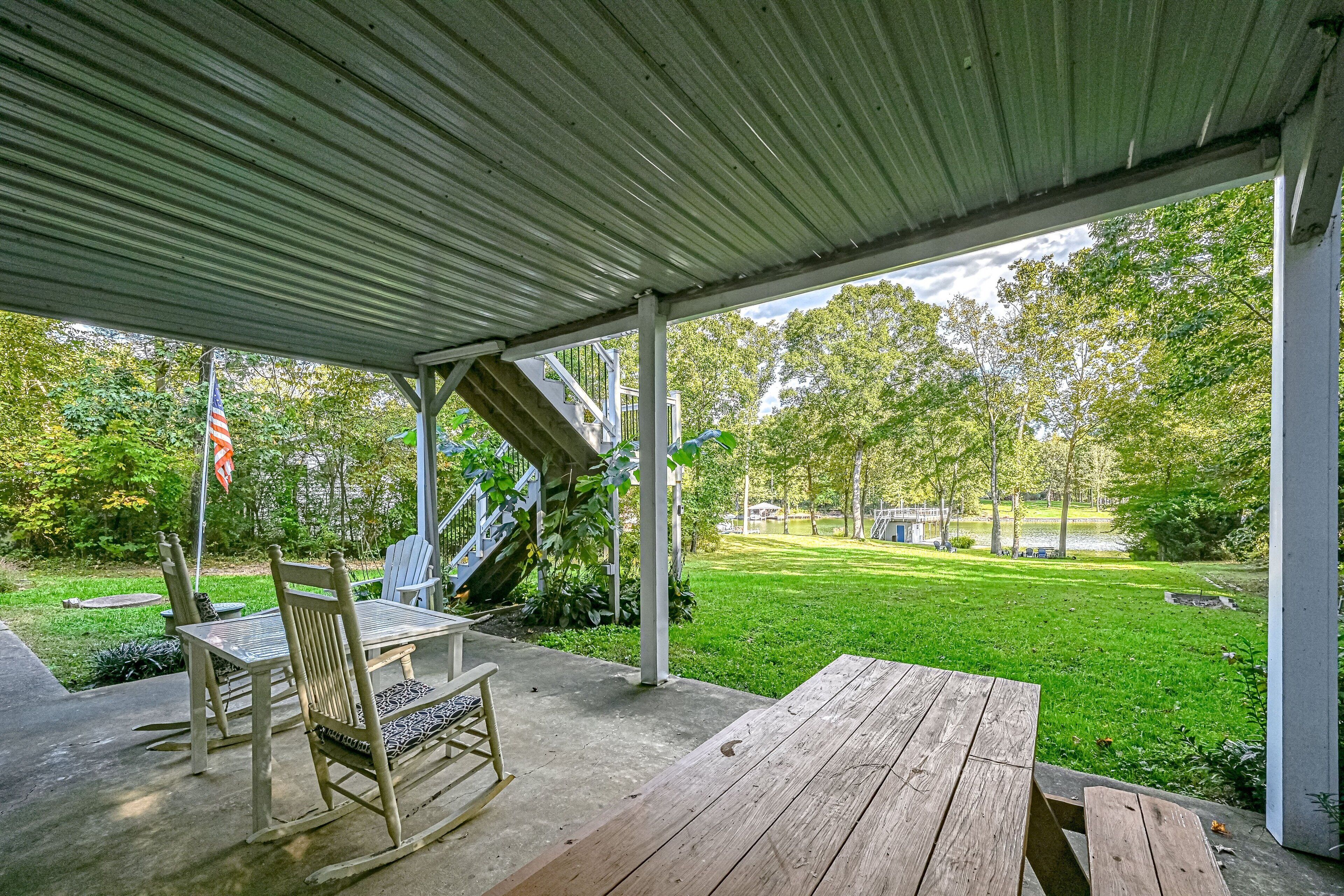 A covered patio with a wooden table and chairs, a flag, and a view of a grassy yard and trees.