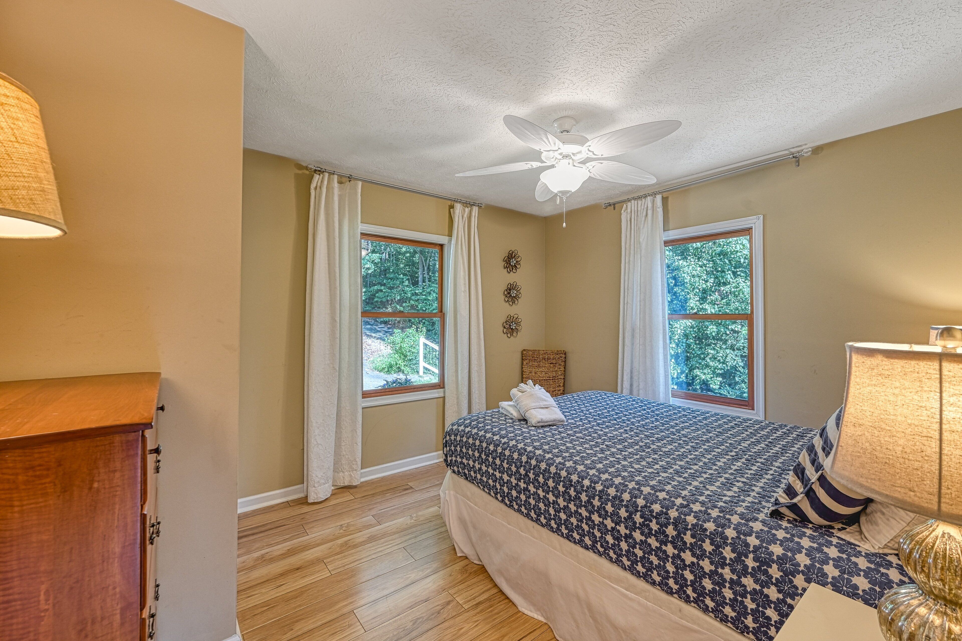 A bedroom with a bed, two windows, a ceiling fan, and a wooden dresser.