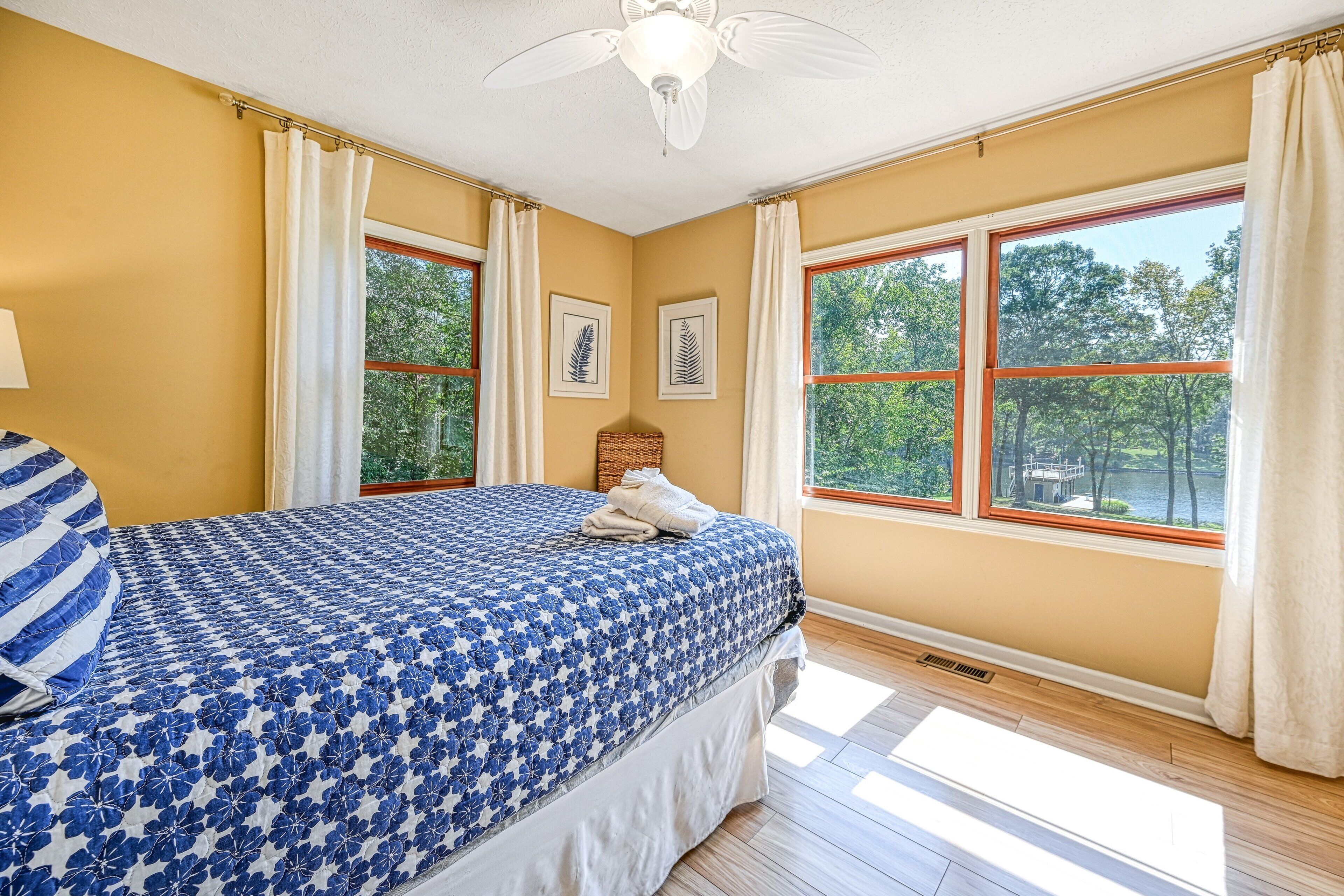 Une chambre à coucher avec un lit, de grandes fenêtres, un ventilateur de plafond et une vue sur les arbres.