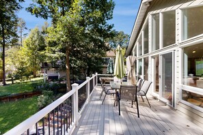 Una terrassa de fusta amb mobiliari exterior, una barana blanca i vista a una zona residencial plena d'arbres i cases.