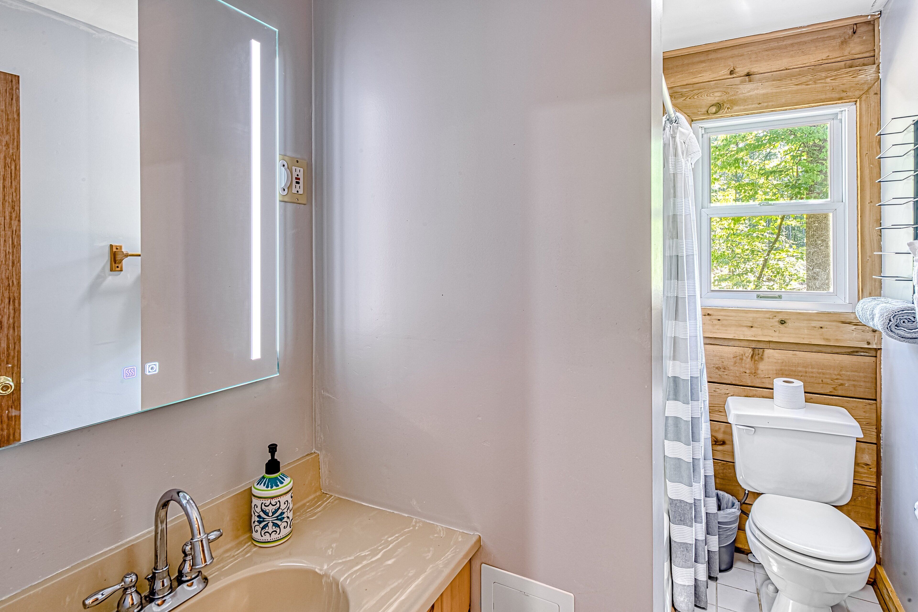 A bathroom with a sink, mirror, toilet, and window.