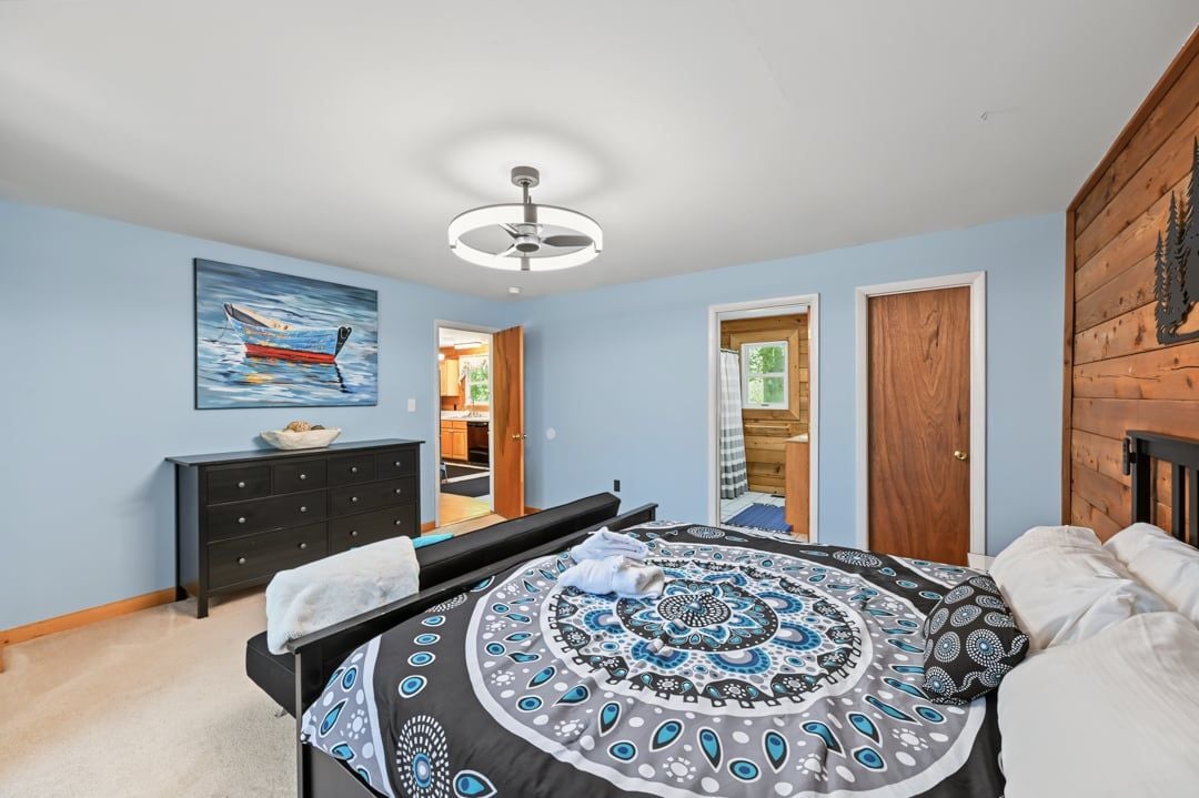 Une chambre à coucher avec un lit, un ventilateur de plafond, un tableau accroché au mur, une commode et une porte en bois.