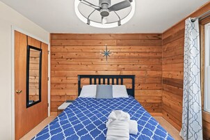 Un dormitorio con cama, ventilador de techo, pared de madera, ventana con cortinas y puerta.