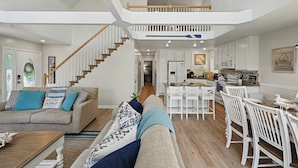Amplia sala de estar con techo alto, una escalera, una cocina con armarios blancos y un comedor con sillas blancas.