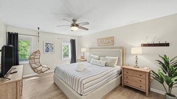 A bedroom with a bed, a ceiling fan, a bedside table with a lamp, a wooden dresser, a chair, and a potted plant.