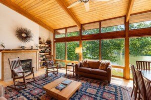 Un salón amplio con chimenea, muebles de madera, ventanas grandes y vistas a un lago.