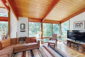 Ein Wohnzimmer mit Holzboden, großen Fenstern, einer Couch, einem Sessel, einem Fernseher und einem Bücherregal.