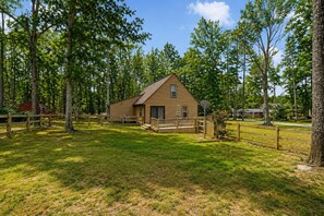 A single-story wooden cabin with a porch, surrounded by a fenced yard and trees. - Lake Anna Home on Large Corner lot With Kayaks, Paddleboard, Firepit & Grill (Bumpass)