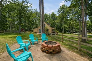 A rustic outdoor seating area with Adirondack chairs, a fire pit, and a wooden cabin in a wooded setting. - Lake Anna Home on Large Corner lot With Kayaks, Paddleboard, Firepit & Grill (Bumpass)