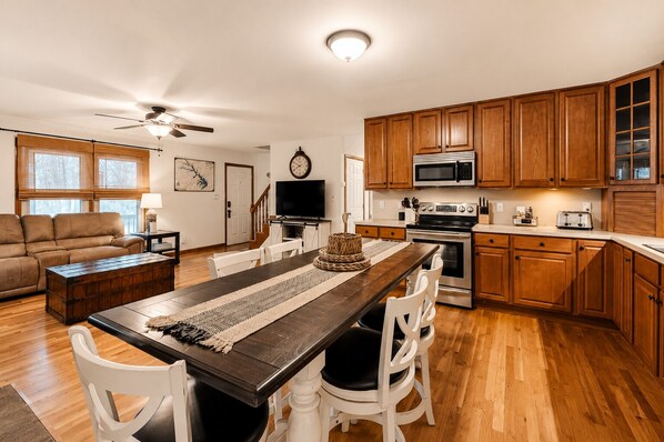 A modern kitchen with wooden cabinets, a microwave, stove, and a dining area with a long table and chairs. - Lake Anna Home on Large Corner lot With Kayaks, Paddleboard, Firepit & Grill (Bumpass)