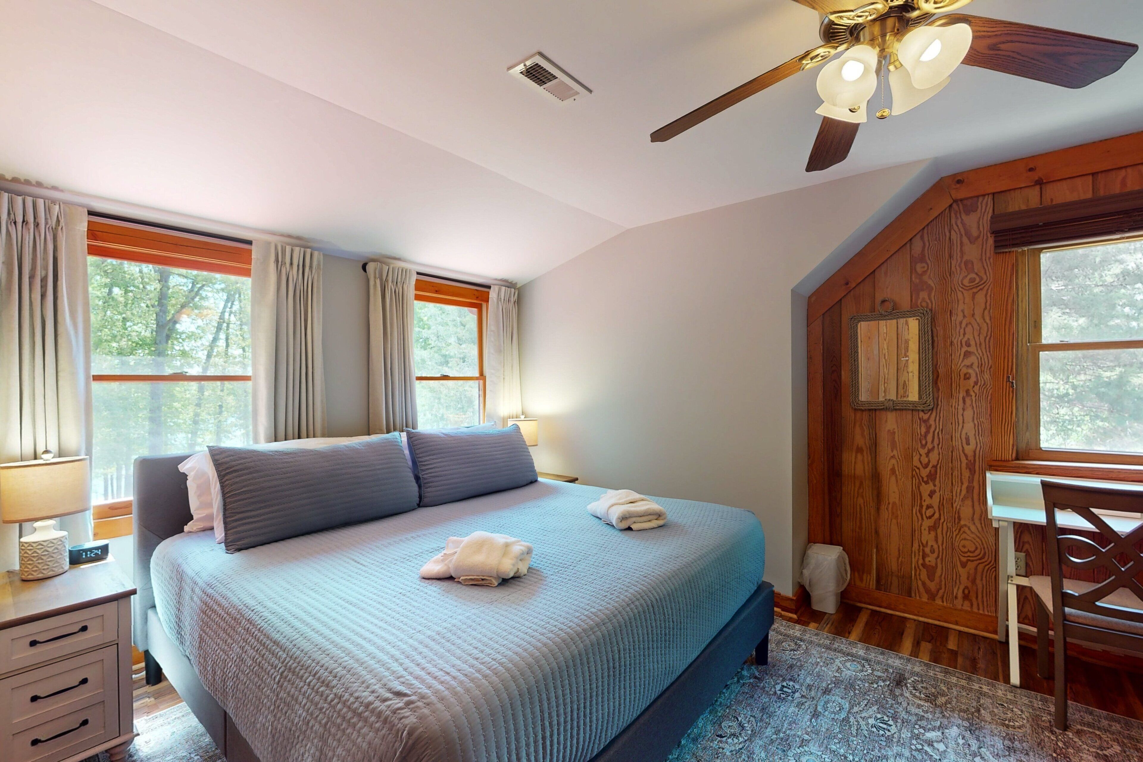 A bedroom with a bed, a desk, a chair, a ceiling fan, and a window with curtains.