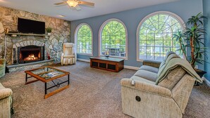 A living room with a stone fireplace, a television, a ceiling fan, and large arched windows. - Lakefront Home With Boat Dock, Private Firepit & Game Room - Dog-friendly (Bumpass)