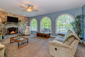 A living room with a stone fireplace, a television, a ceiling fan, and large arched windows. - Lakefront Home With Boat Dock, Private Firepit & Game Room - Dog-friendly (Bumpass)