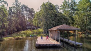 A dock with chairs, a covered area, and a house in a wooded area. - Lakefront Home With Boat Dock, Private Firepit & Game Room - Dog-friendly (Bumpass)