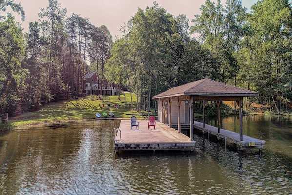 A dock with chairs, a covered area, and a house in a wooded area. - Lakefront Home With Boat Dock, Private Firepit & Game Room - Dog-friendly (Bumpass)
