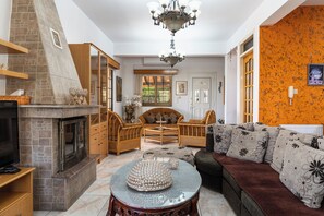 Un salon dotĂ© dâune cheminĂ©e, dâun tĂ©lĂ©viseur, dâun coin repas avec des meubles en osier et dâun canapĂ© agrĂ©mentĂ© de coussins Ă motifs floraux.