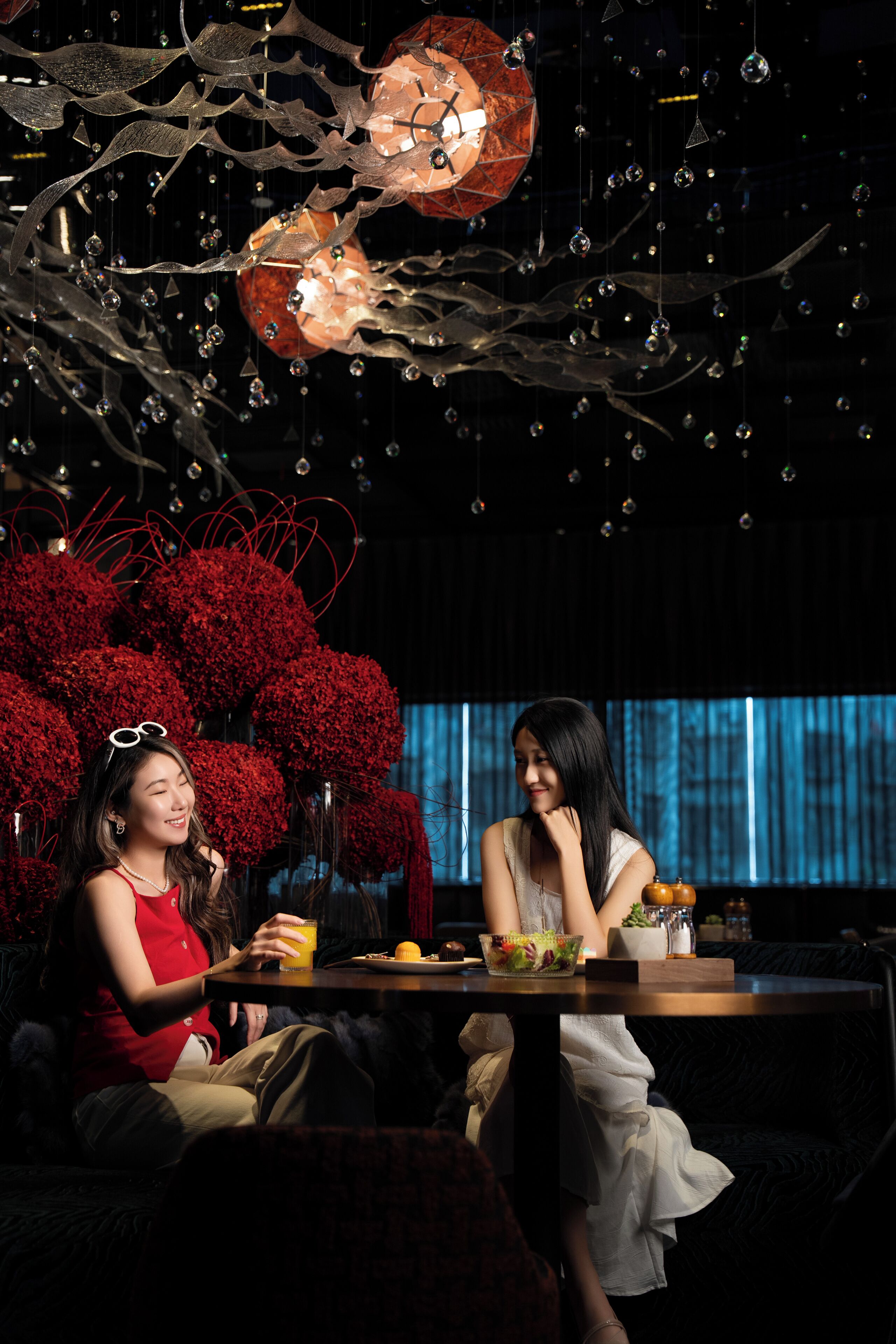 Twee personen zitten aan een tafel met een drankje en een fruitschaal, omgeven door decoratieve objecten en een rood bloemstuk.