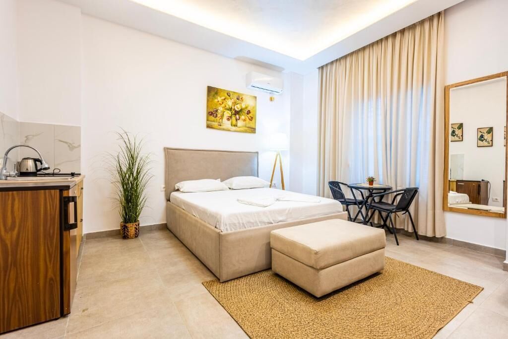 A modern hotel room with a bed, a desk, a chair, a small table, a potted plant, and a painting on the wall.