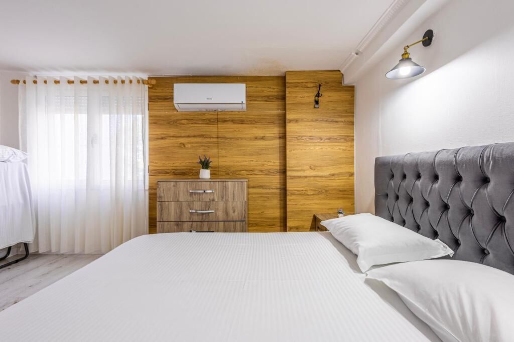 A bedroom with a bed, a wooden dresser, a wall-mounted air conditioner, and a bedside lamp.