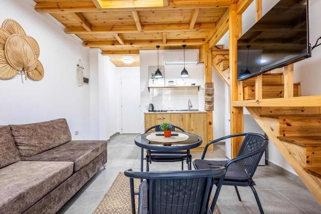 A modern living room with a sofa, a round table, two chairs, a TV, wooden beams, and a kitchen in the background.