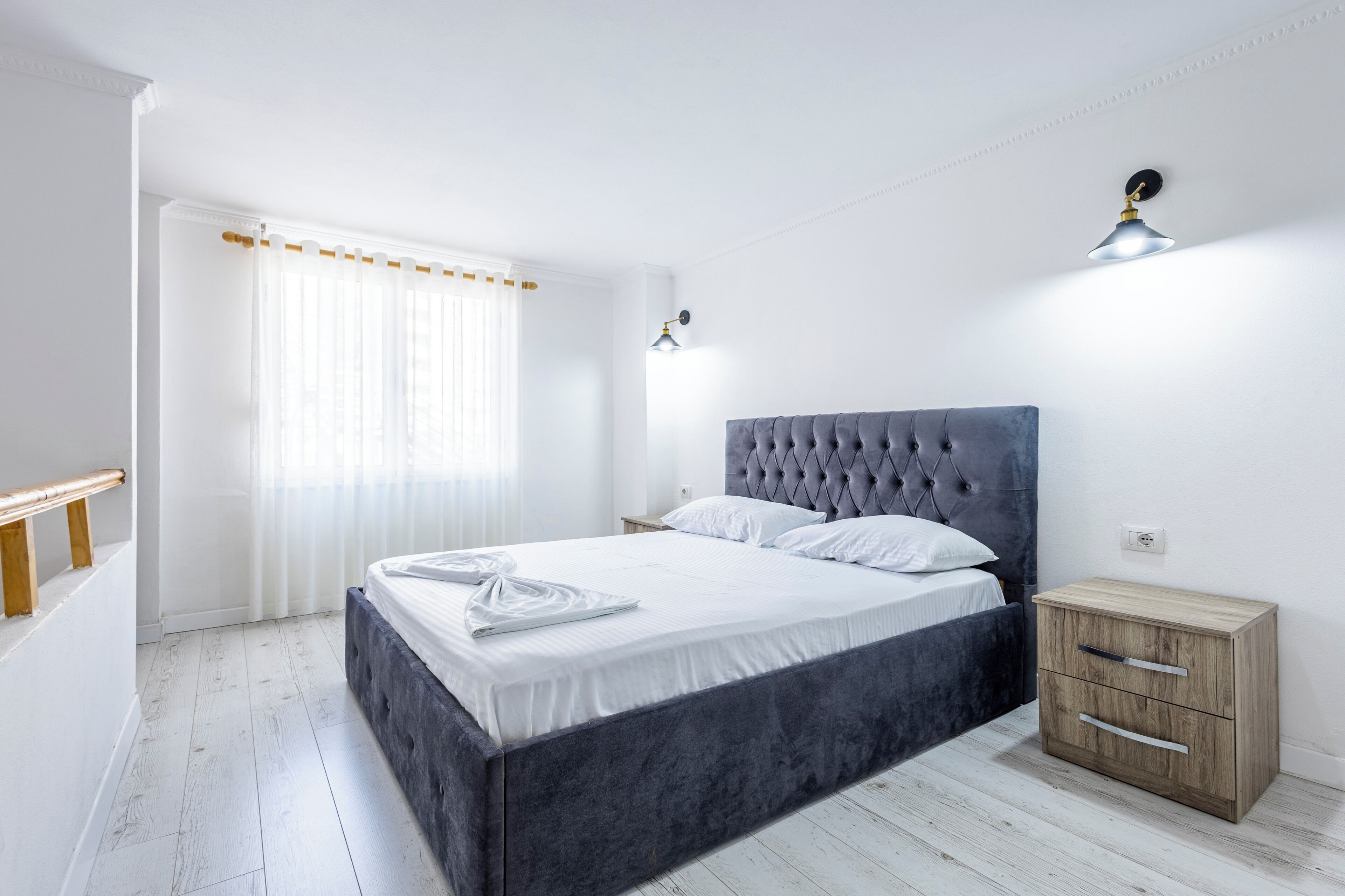 A bedroom with a bed, bedside tables, a nightstand, a window with curtains, and a wall-mounted light fixture.