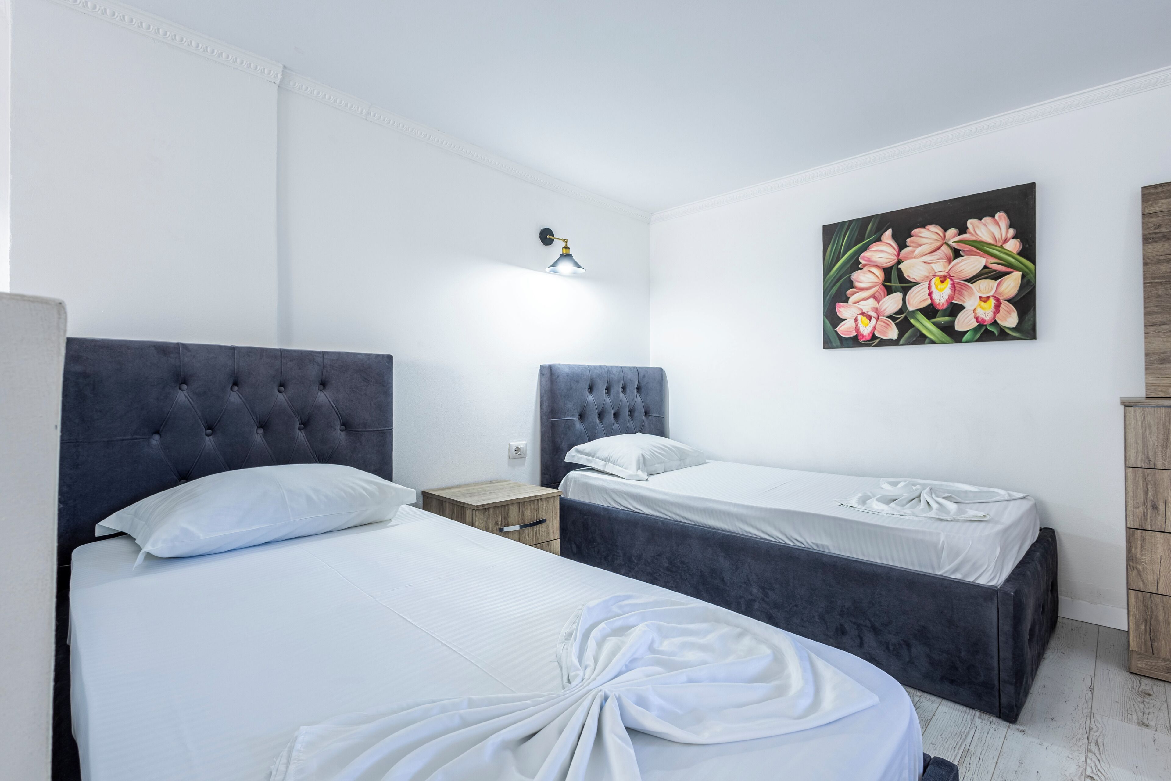 A bedroom with two beds, a nightstand, a wall-mounted lamp, and a floral painting.