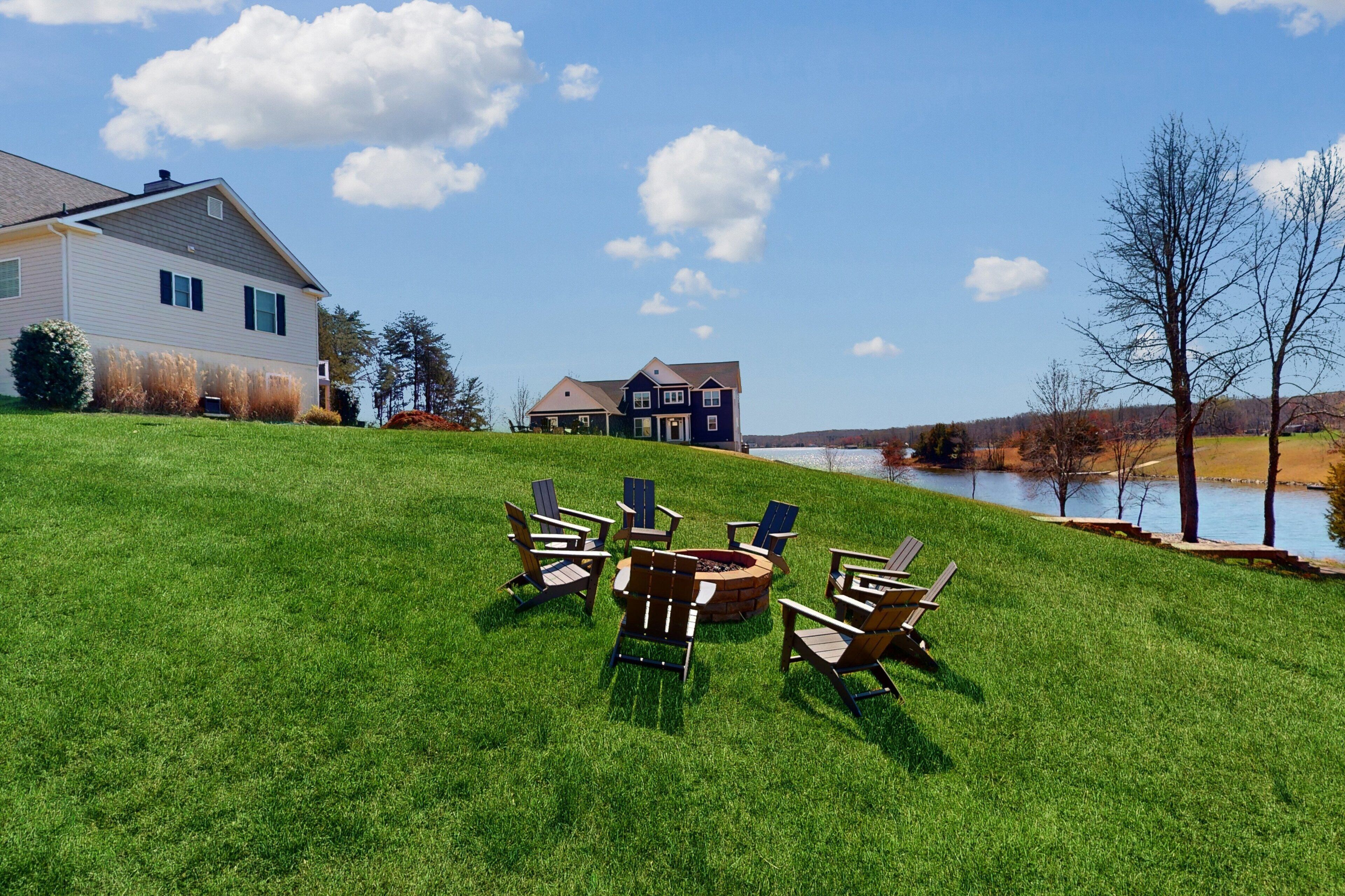 A grassy area with outdoor seating and a fire pit, a lake, and two houses in the background.