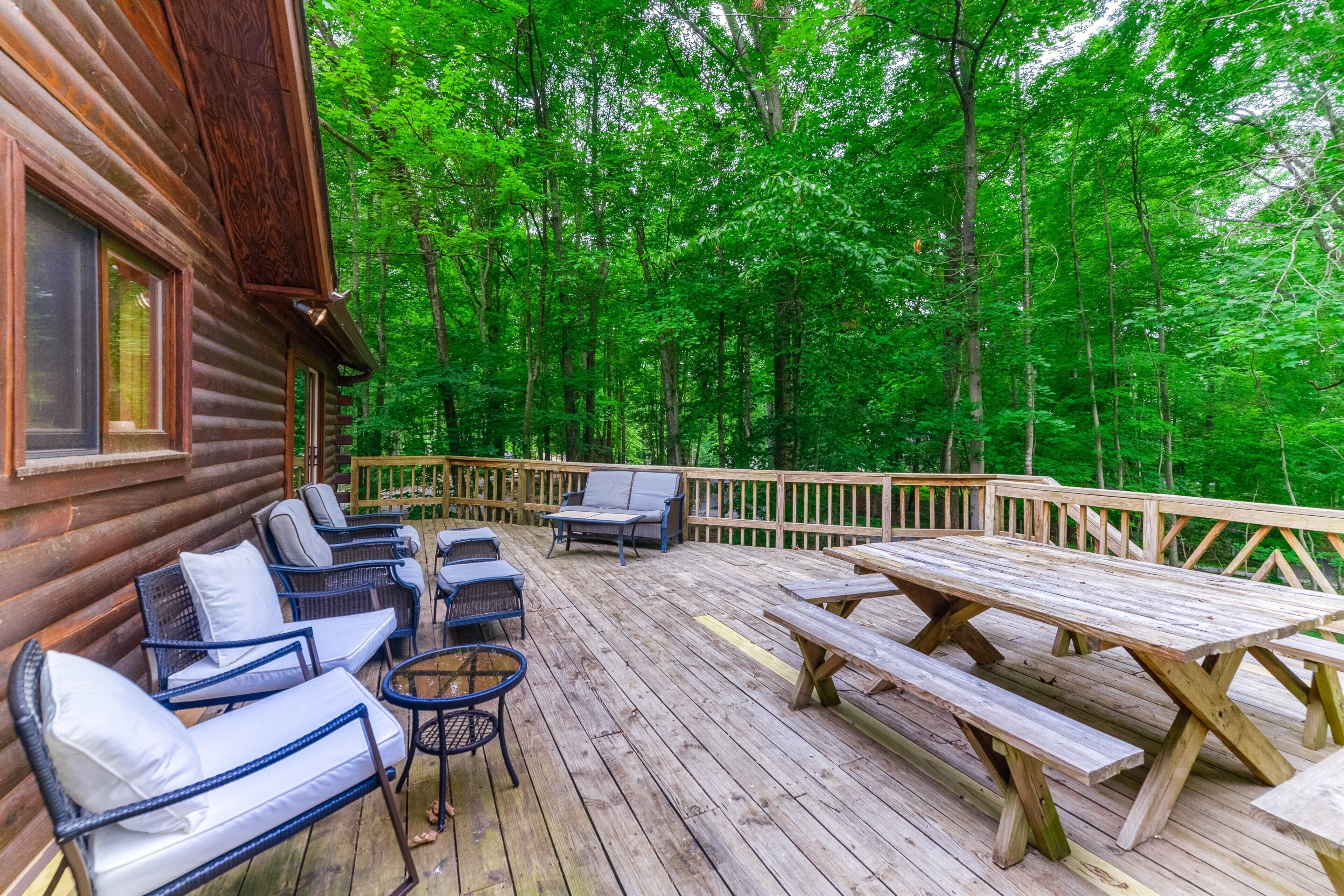 Una terraza de madera con mobiliario exterior, incluyendo sofás y una mesa, rodeada por un bosque.
