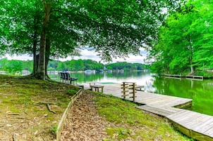 A lakeside park with a wooden dock, benches, and a tree providing shade.