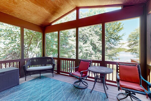 A covered outdoor area with a wooden ceiling, large windows, a wicker sofa, a red chair, and a small round table.