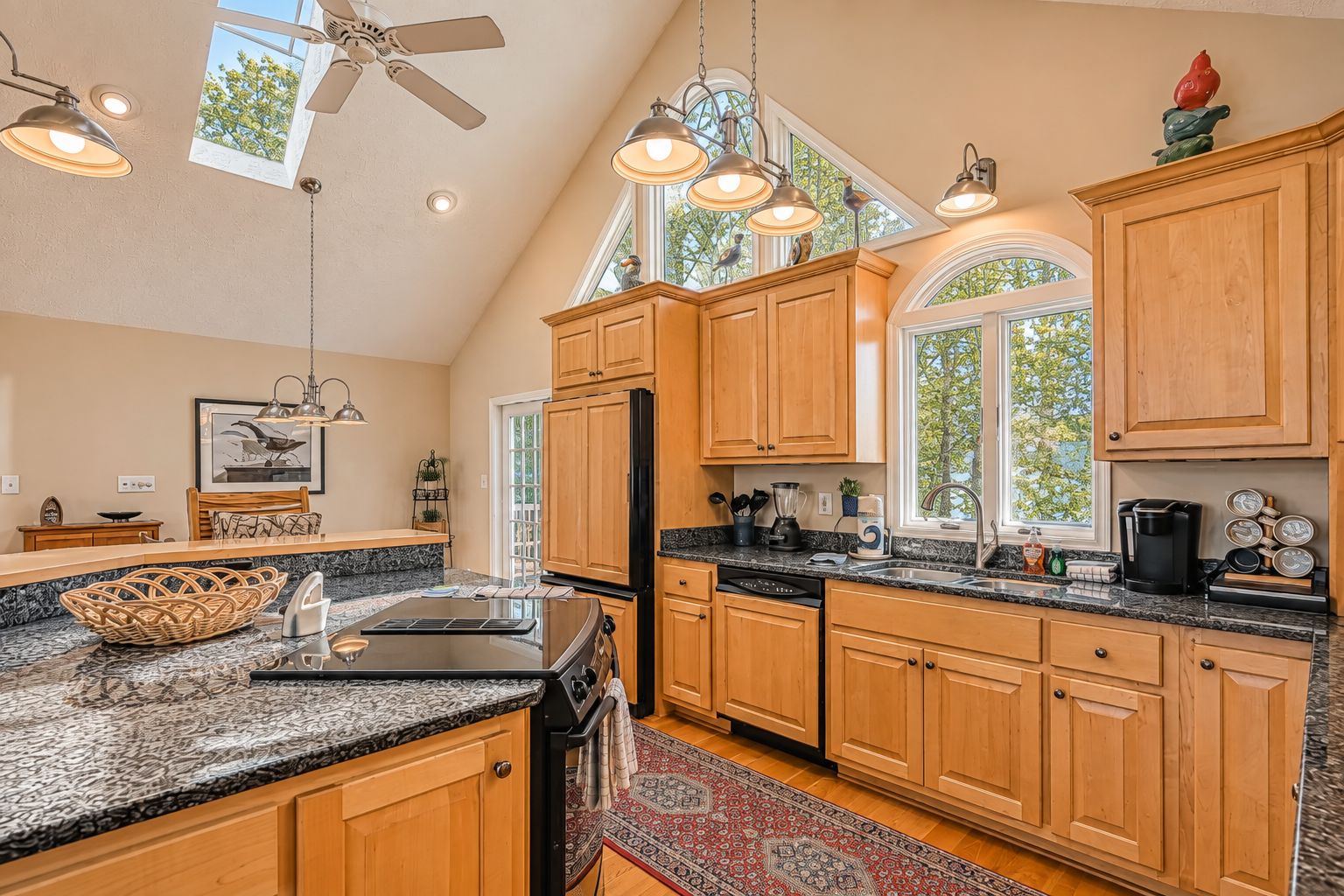 A kitchen with wooden cabinets, granite countertops, a black refrigerator, and a window with a view of trees.