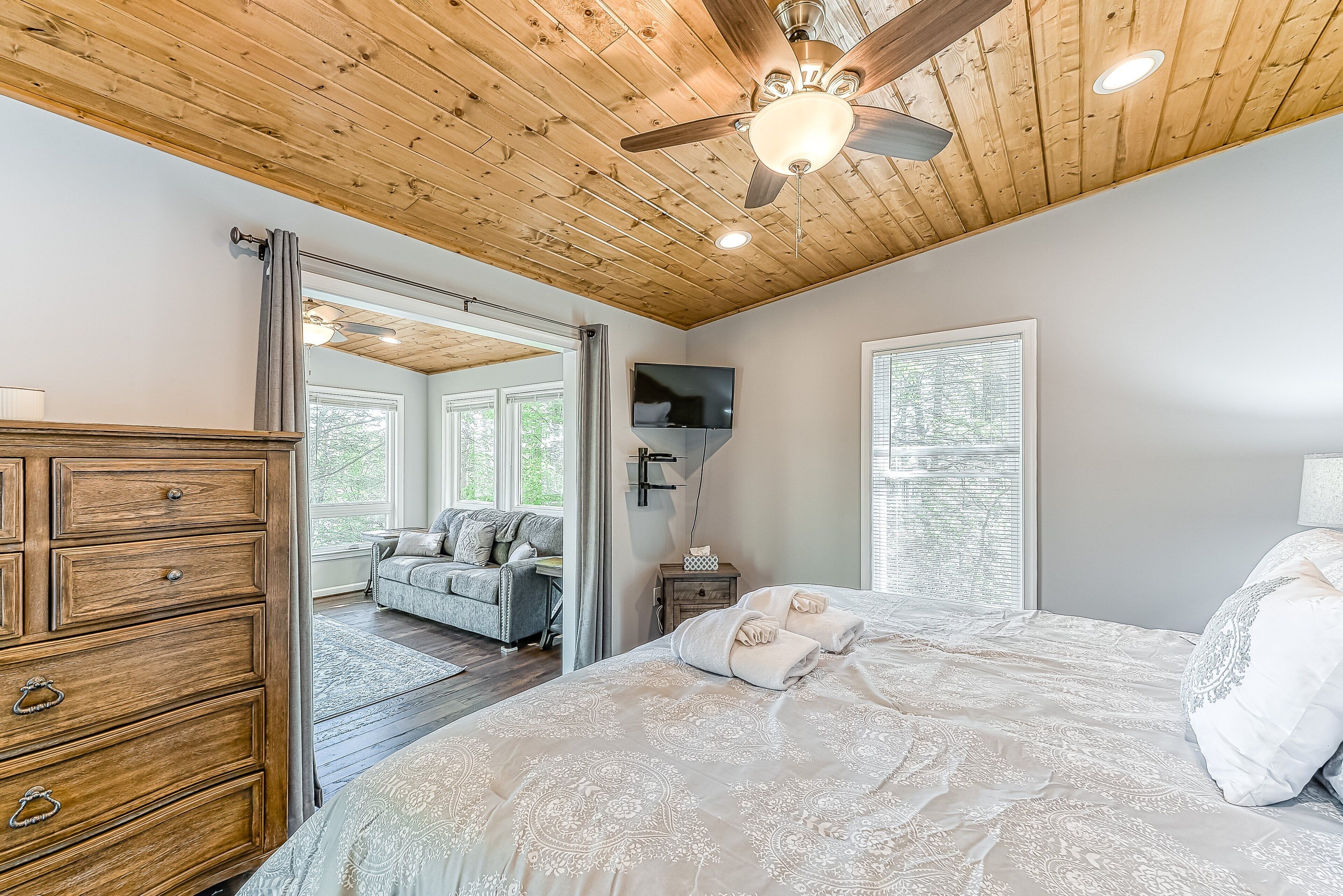 Une chambre avec un plafond en bois, un lit recouvert d’une couvre-lit à motifs, une commode, un téléviseur et une fenêtre avec des stores.