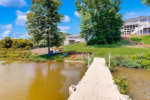 A riverside dock with a boat, a house with a balcony, and a grassy area. - Two-story, Lakefront Getaway With Dock, Beach, Grill, 2 Kayaks, 2 Canoes, AC (Orange)