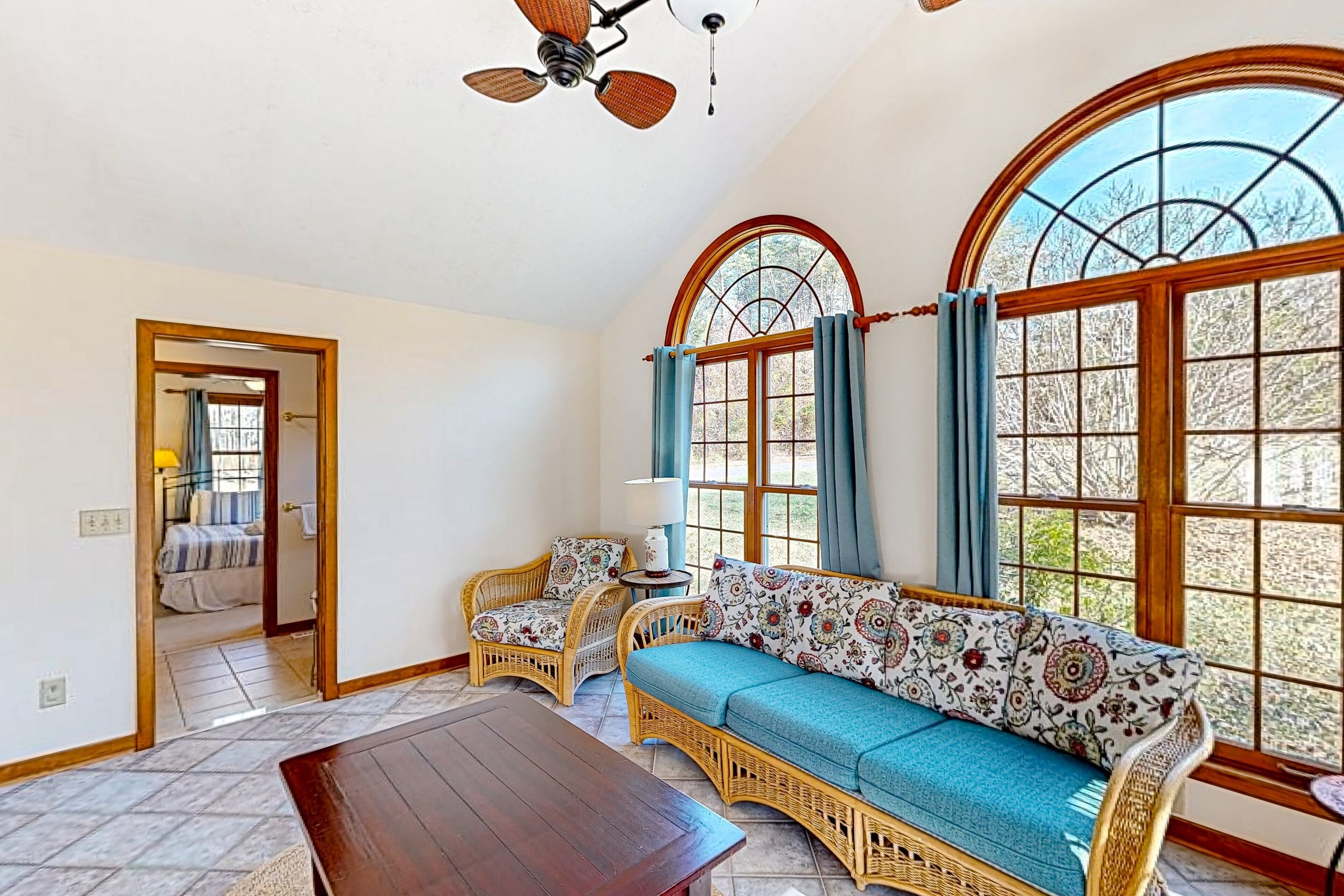 A room with a wooden table, wicker sofa, and chairs, large arched windows, and a ceiling fan.